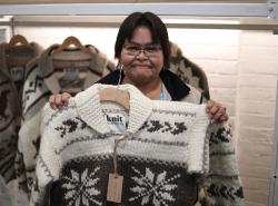 A woman with short dark hair and wearing a Cowichan sweater holds up a Cowichan sweater with a snowflake design in front of her. Behind her hang Cowichan sweaters.
