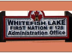 Two wooden posts hold up a sign with a red maple leaf with a tipi overlaid on it. The sign reads WHITEFISH LAKE FIRST NATIONS #128 Administration Office