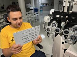 A man sits in an optometry office. He holds a vision test card in front of him.