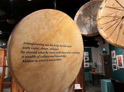 Indigenous hide hand drums hang in a grouping and there are verses of a poem printed on each of them.