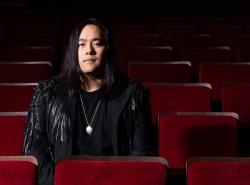 A man with long black hair sits alone in a theatre with red velvet chairs.