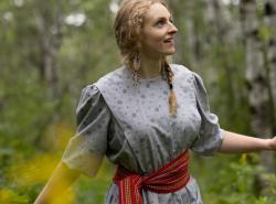 A young girl in a cotton dress with a Metis sash around her waist walks through the woods.