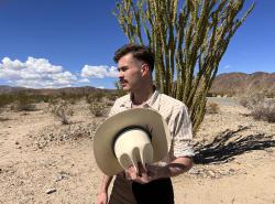 A man stands with a cowboy hat in his hands while out in a desert-like landscape.