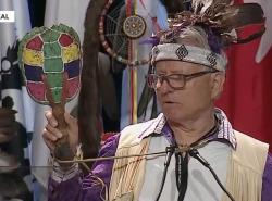 A man in a beaded headdress with feathers holds up a painted turtle ratte as he speaks at a microphone.